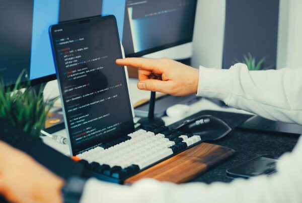 A developer pointing at code on a laptop screen at a multi-monitor workstation, representing editing Shopify theme code to fix product URL structure for SEO.