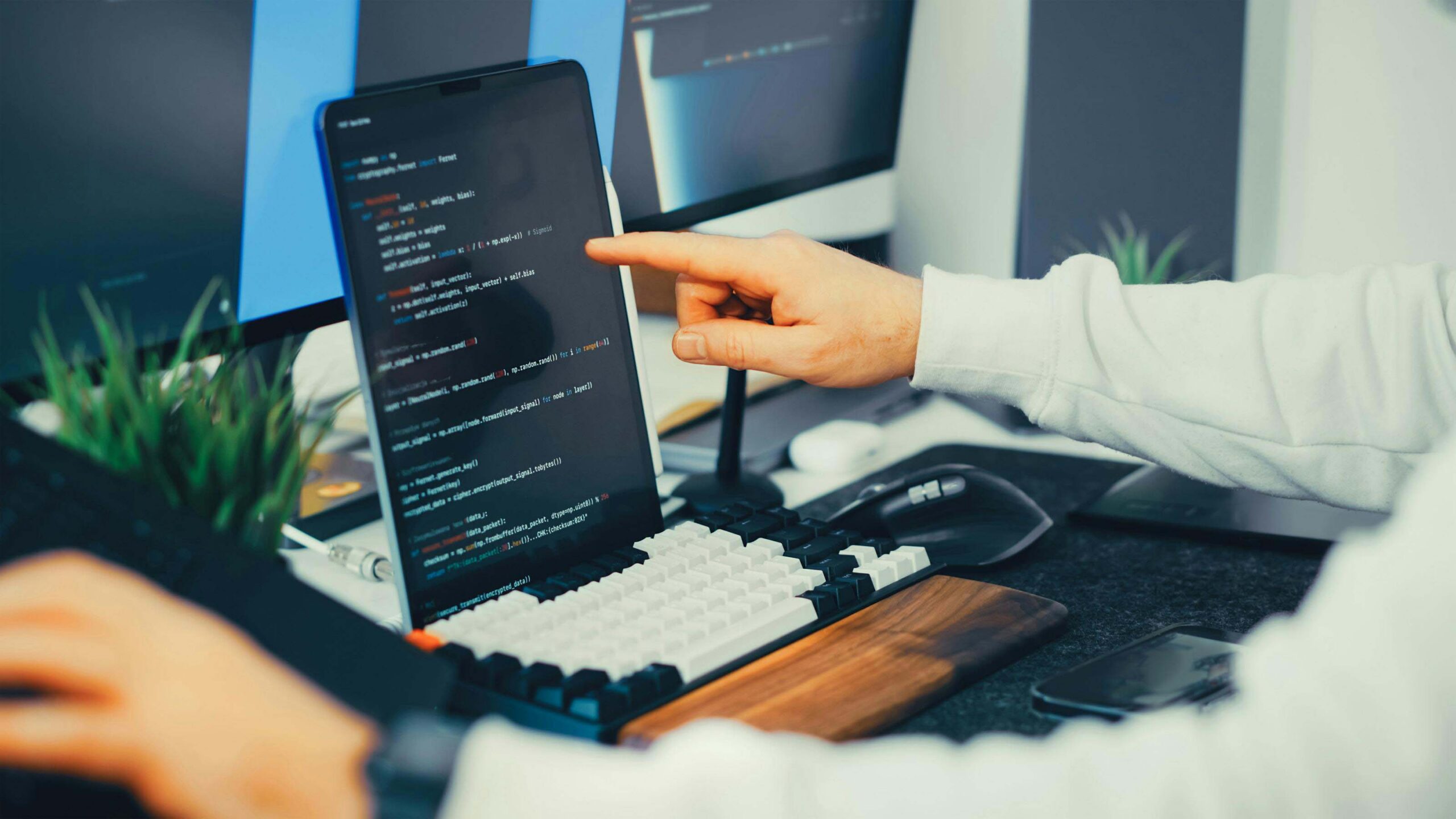 A developer pointing at code on a laptop screen at a multi-monitor workstation, representing editing Shopify theme code to fix product URL structure for SEO.